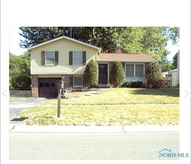 split level home with a garage, driveway, a front lawn, brick siding, and a chimney