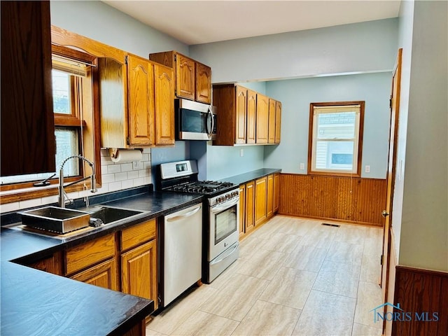 kitchen with appliances with stainless steel finishes, a sink, brown cabinets, wainscoting, and dark countertops