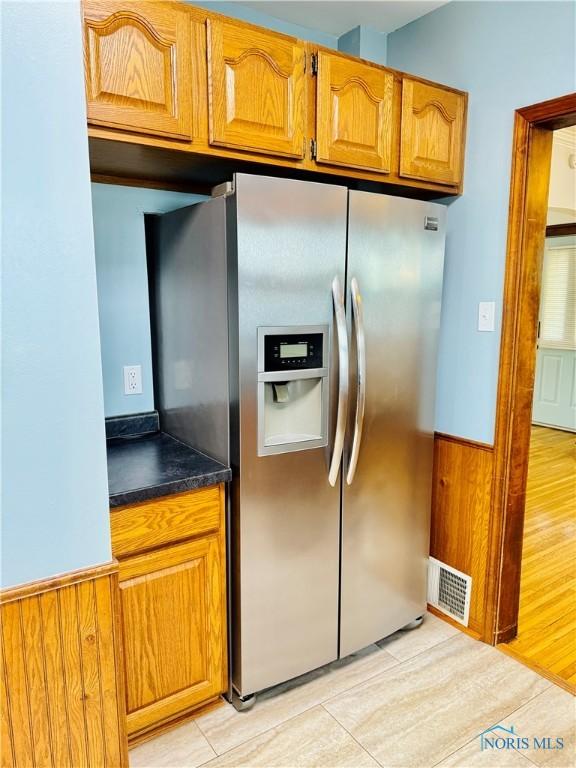 kitchen with stainless steel fridge, dark countertops, brown cabinetry, wainscoting, and light wood finished floors