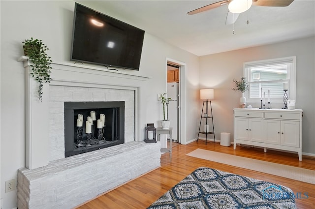 living area with a ceiling fan, a stone fireplace, light wood-type flooring, and baseboards