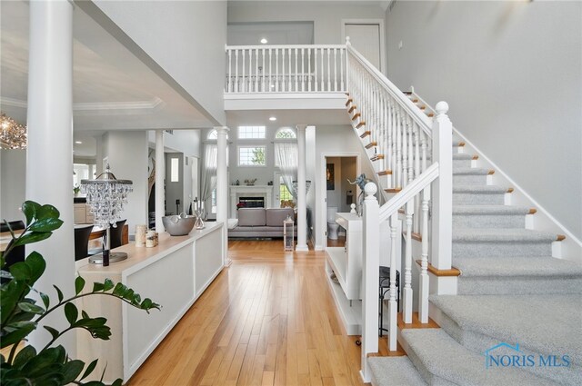 entrance foyer with ornate columns, a glass covered fireplace, stairs, light wood-style floors, and a towering ceiling