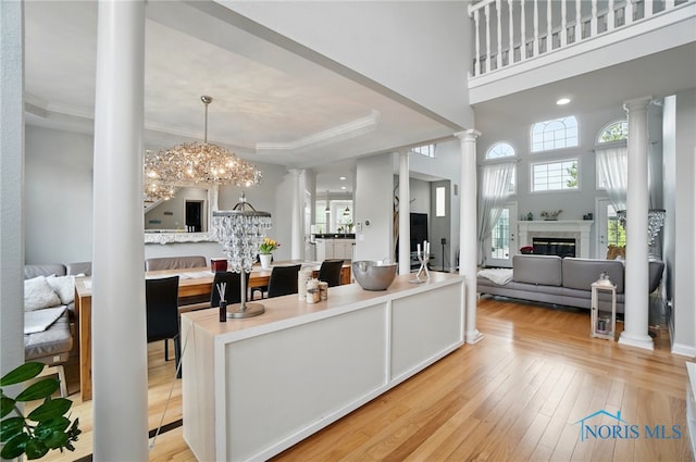 living area featuring ornate columns, light wood-style floors, a glass covered fireplace, crown molding, and a chandelier