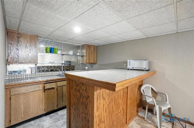 4871 Olde Meadow Ln, Sylvania, OH 43560 - kitchen featuring tile counters, a peninsula, a drop ceiling, a kitchen breakfast bar, and brown cabinets
