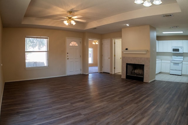 unfurnished living room featuring a tiled fireplace, ceiling fan, dark hardwood / wood-style flooring, and a tray ceiling