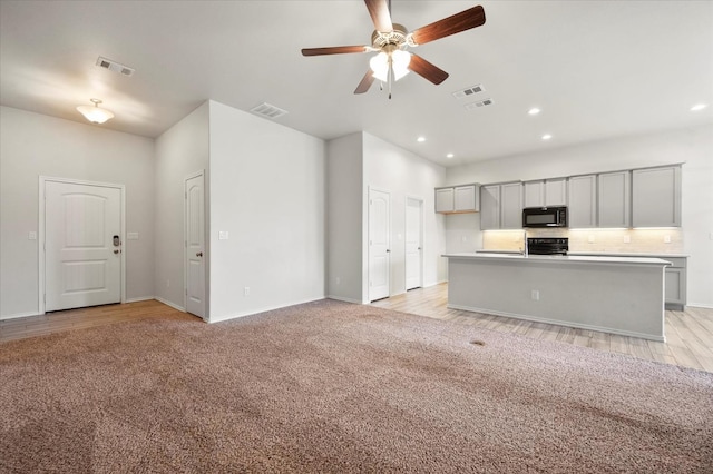 kitchen with light carpet, gray cabinets, an island with sink, ceiling fan, and black appliances