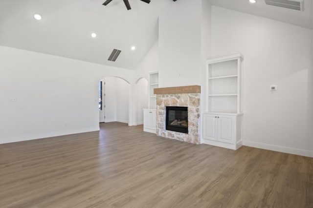 unfurnished living room with ceiling fan, hardwood / wood-style floors, high vaulted ceiling, built in shelves, and a stone fireplace