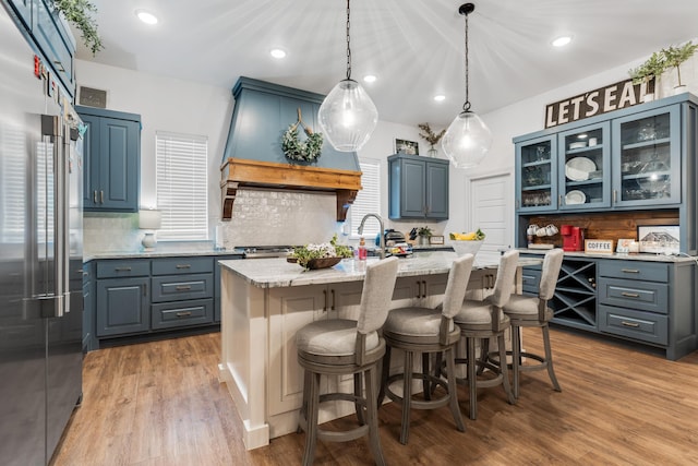 kitchen with glass insert cabinets, high end refrigerator, light wood-style floors, hanging light fixtures, and a center island with sink