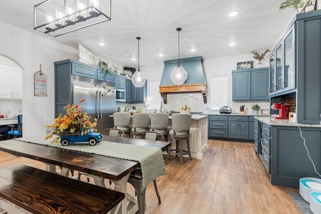 kitchen with glass insert cabinets, decorative light fixtures, stainless steel built in fridge, blue cabinetry, and premium range hood