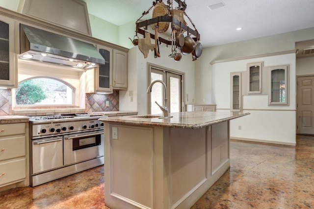 kitchen with wall chimney range hood, tasteful backsplash, an island with sink, french doors, and range with two ovens