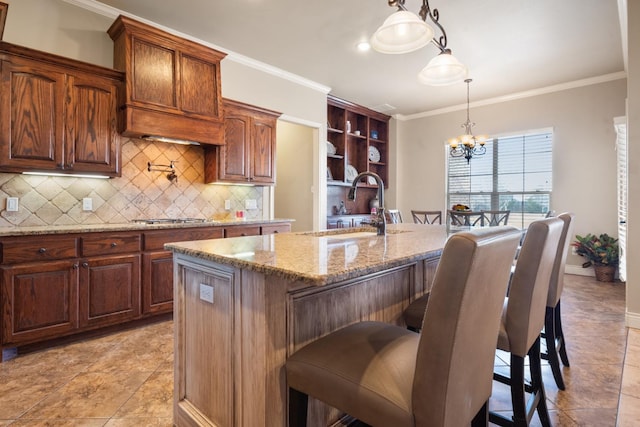 kitchen featuring a kitchen island with sink, a sink, crown molding, decorative backsplash, and a chandelier