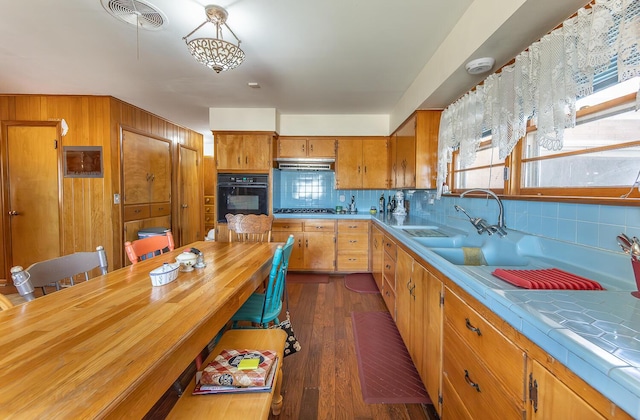 kitchen with brown cabinetry, black oven, tile countertops, under cabinet range hood, and backsplash