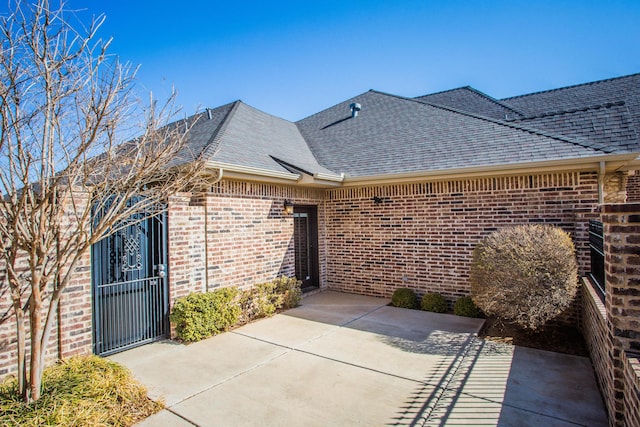 view of exterior entry with a shingled roof, a patio area, a gate, and brick siding