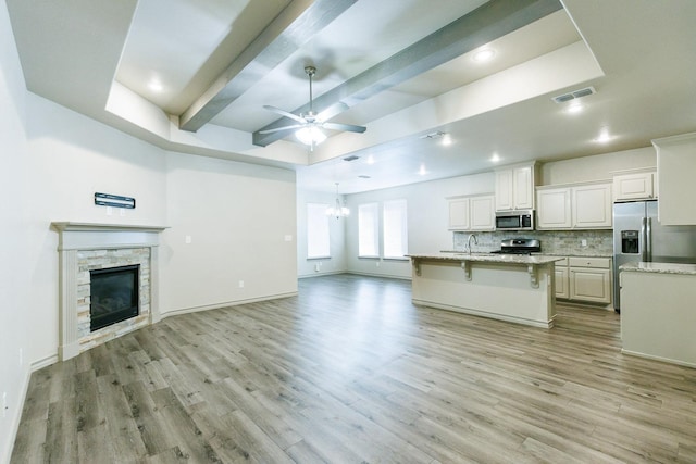 unfurnished living room with beamed ceiling, a stone fireplace, ceiling fan with notable chandelier, and light wood-type flooring