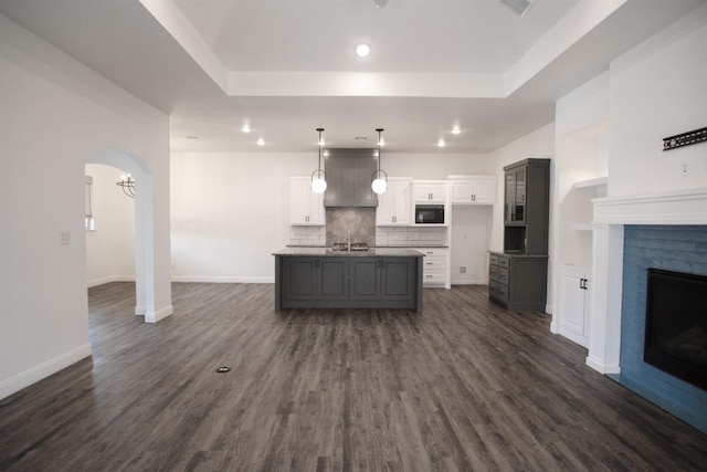 kitchen with black microwave, white cabinets, decorative backsplash, dark wood-type flooring, and a center island with sink