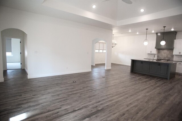 unfurnished living room featuring a raised ceiling, sink, dark wood-type flooring, and ceiling fan with notable chandelier