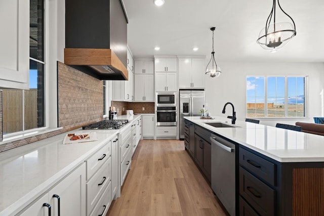 kitchen featuring decorative light fixtures, ventilation hood, white cabinets, stainless steel appliances, and a center island with sink