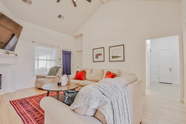 living room with ceiling fan, a tiled fireplace, high vaulted ceiling, and light wood-type flooring