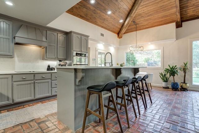 kitchen with premium range hood, wood ceiling, stainless steel oven, beam ceiling, and a kitchen island with sink