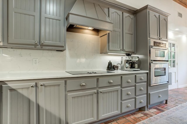 kitchen featuring premium range hood, black electric cooktop, gray cabinets, stainless steel double oven, and decorative backsplash