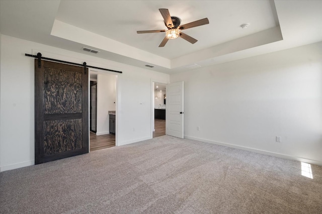 unfurnished bedroom with a tray ceiling, a barn door, ceiling fan, and carpet flooring