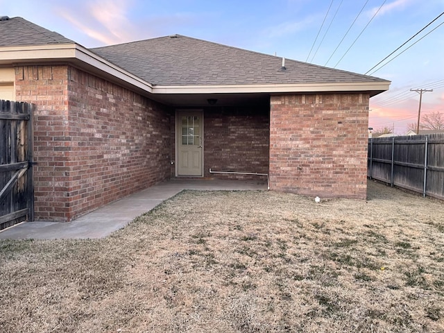 exterior entry at dusk with roof with shingles, fence, and brick siding