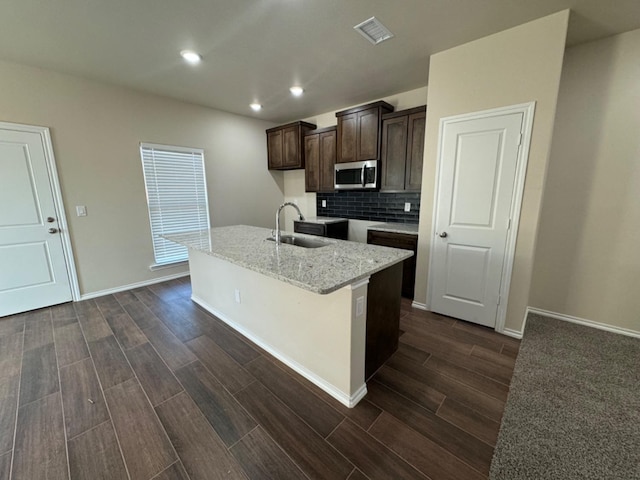 kitchen featuring sink, backsplash, dark brown cabinetry, light stone countertops, and an island with sink