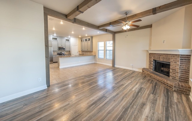 unfurnished living room featuring a brick fireplace, hardwood / wood-style flooring, beamed ceiling, and ceiling fan