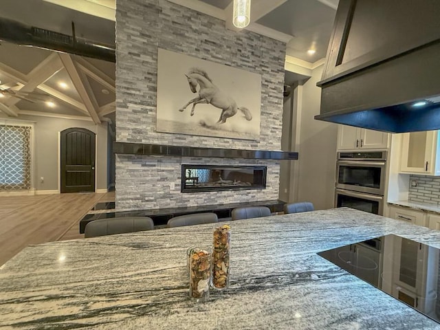 kitchen featuring beam ceiling, light stone counters, a fireplace, white cabinets, and stainless steel double oven