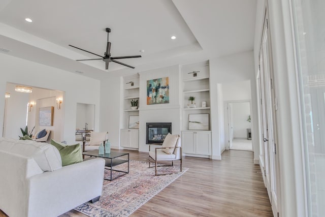 living room with a raised ceiling, ceiling fan with notable chandelier, light wood-type flooring, and built in shelves