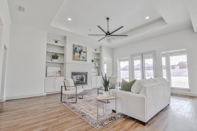 living room with a raised ceiling, ceiling fan, built in features, and light wood-type flooring