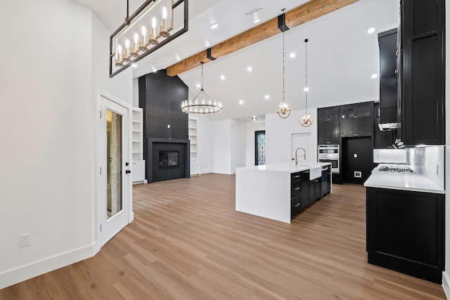 kitchen featuring a fireplace, hanging light fixtures, a kitchen island with sink, beam ceiling, and light hardwood / wood-style floors