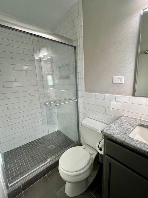 bathroom featuring a shower with shower door, tile walls, tile patterned flooring, vanity, and toilet