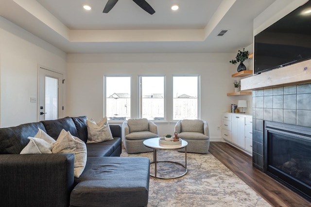 living area with ceiling fan, a tiled fireplace, a tray ceiling, and dark wood finished floors