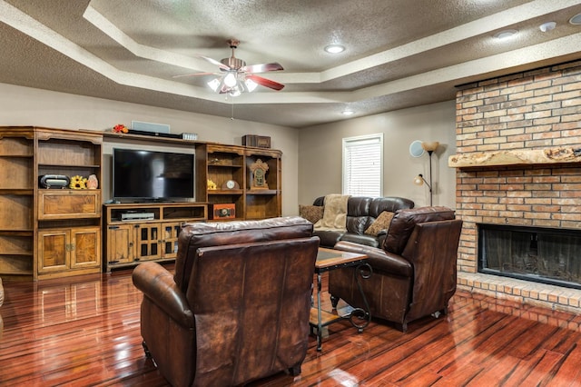 living room featuring dark hardwood / wood-style floors, ceiling fan, a tray ceiling, a brick fireplace, and a textured ceiling