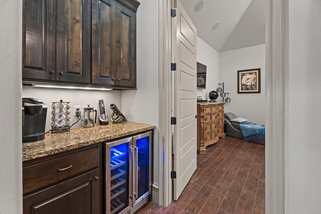 bar with vaulted ceiling, dark brown cabinets, wine cooler, and light stone counters