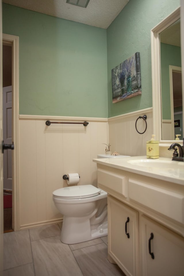 bathroom featuring a decorative wall, a textured ceiling, vanity, toilet, and wainscoting