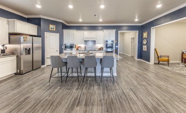 kitchen featuring white cabinetry, stainless steel appliances, light stone counters, an island with sink, and light wood-type flooring