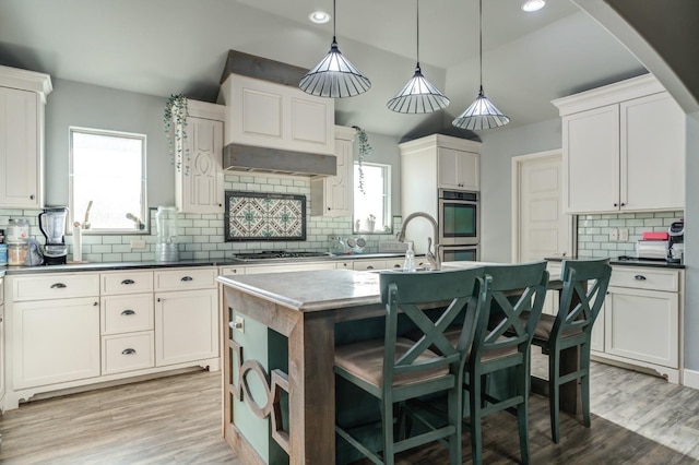 kitchen featuring pendant lighting, stainless steel appliances, ventilation hood, an island with sink, and white cabinets