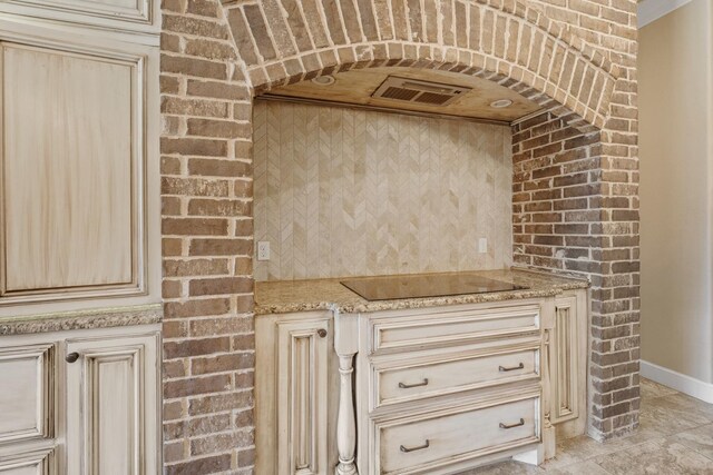 kitchen featuring light stone counters, brick wall, black electric stovetop, and cream cabinets