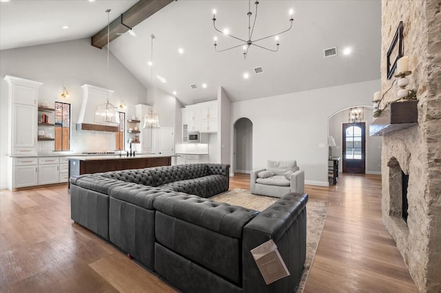 living room with beam ceiling, high vaulted ceiling, a notable chandelier, a stone fireplace, and light wood-type flooring