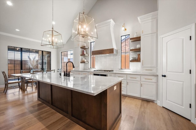 kitchen featuring pendant lighting, premium range hood, white cabinetry, a kitchen island with sink, and light stone countertops