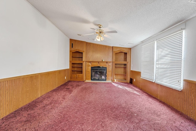 unfurnished living room featuring a textured ceiling, wooden walls, built in features, ceiling fan, and carpet