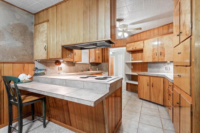 kitchen featuring a breakfast bar area, ceiling fan, light tile patterned flooring, kitchen peninsula, and wood walls