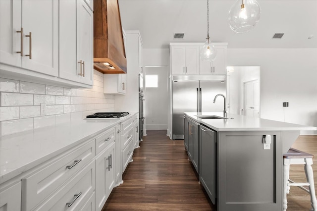 kitchen featuring custom exhaust hood, white cabinetry, decorative light fixtures, an island with sink, and stainless steel appliances