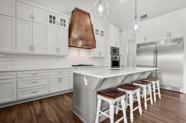 kitchen featuring a center island with sink, built in appliances, custom exhaust hood, and white cabinets