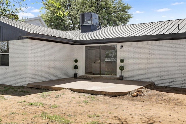 doorway to property featuring a patio, central AC unit, a chimney, brick siding, and metal roof