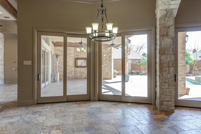 doorway to outside featuring stone tile floors, ornate columns, crown molding, baseboards, and ceiling fan with notable chandelier