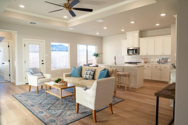 living room with sink, light wood-type flooring, ornamental molding, a tray ceiling, and ceiling fan