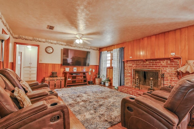 living room with ceiling fan, a brick fireplace, wooden walls, and a textured ceiling