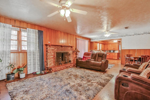 living room featuring a brick fireplace, a textured ceiling, wooden walls, ceiling fan, and light hardwood / wood-style floors